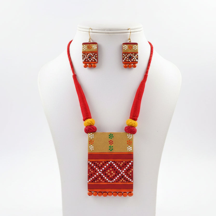 A red and orange geometric patterned necklace set displayed on a black mannequin bust, with matching earrings on black stands, placed on artificial green grass.