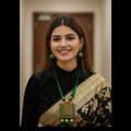 A woman with long brown hair smiling, wearing a black and gold saree with a prominent green and gold pendant necklace and matching green earrings, set against a softly blurred indoor background.