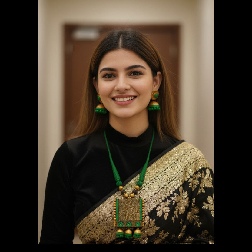 A woman with long brown hair smiling, wearing a black and gold saree with a prominent green and gold pendant necklace and matching green earrings, set against a softly blurred indoor background.