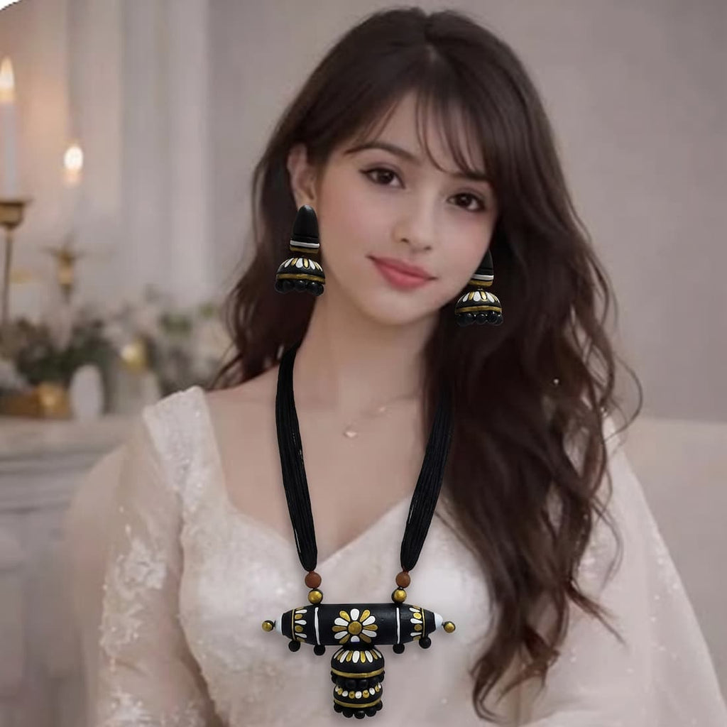 A woman seated indoors wearing a white embroidered traditional outfit with a black-and-gold statement necklace, matching drop earrings, and stacked gold bangles.