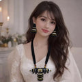 A woman seated indoors wearing a white embroidered traditional outfit with a black-and-gold statement necklace, matching drop earrings, and stacked gold bangles.