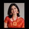 A woman with dark shoulder-length hair smiling, wearing a red and gold patterned traditional sari, a matching red necklace with a large square gold-toned pendant, and matching red and gold earrings, set against a plain light gray background.
