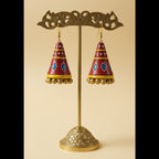 A pair of handmade red, yellow, and blue jhumka earrings displayed on a white rectangular stand.