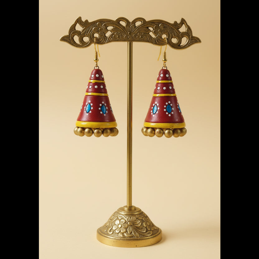 A pair of handmade red, yellow, and blue jhumka earrings displayed on a white rectangular stand.