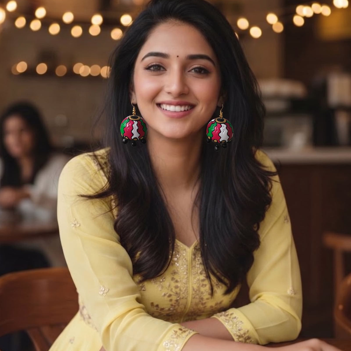 A smiling woman with long dark hair wearing a light yellow embroidered kurta and large colorful jhumka earrings, seated at a table in a warmly lit cafe or restaurant.