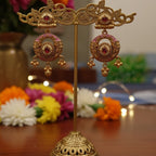 A pair of ornate gold-toned earrings with pink enamel and red stone accents, displayed on a decorative gold stand over a wooden table with blurred flowers in the background.