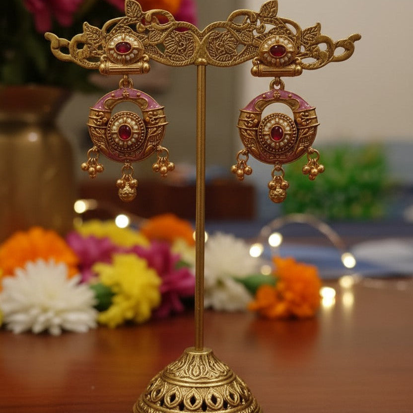 A pair of ornate gold-toned earrings with pink enamel and red stone accents, displayed on a decorative gold stand over a wooden table with blurred flowers in the background.