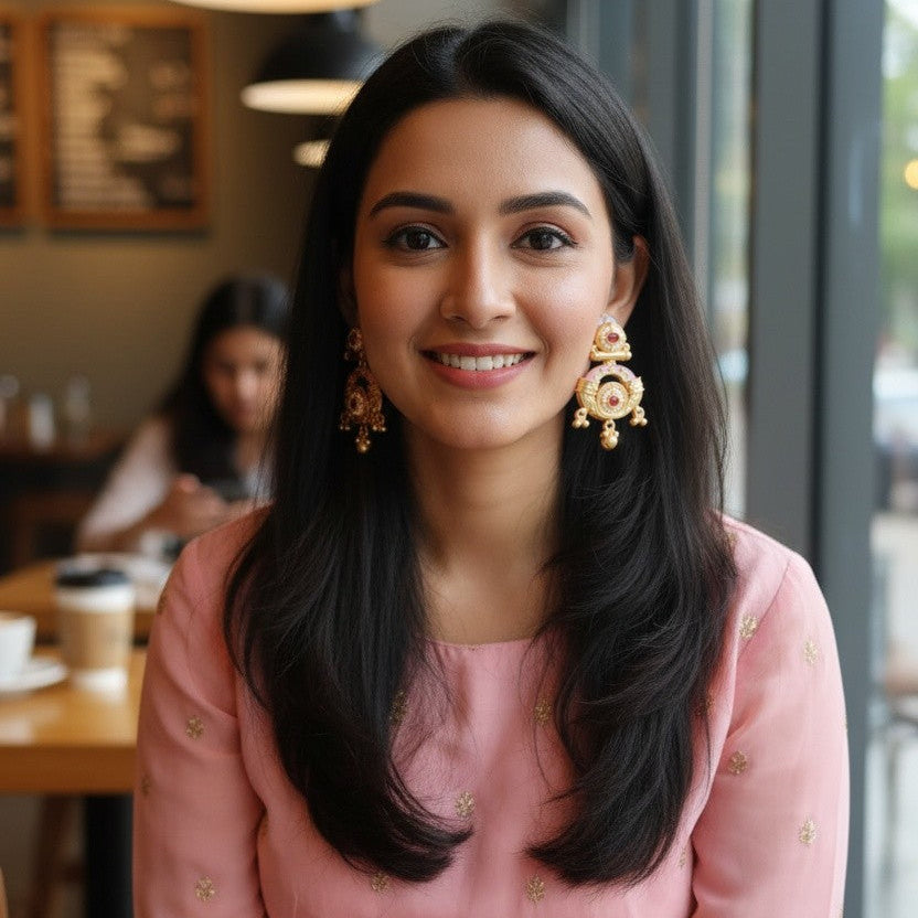 A woman with long black hair is seated in a cafe setting, wearing a light pink traditional-style dress with small gold motifs and large ornate gold earrings.