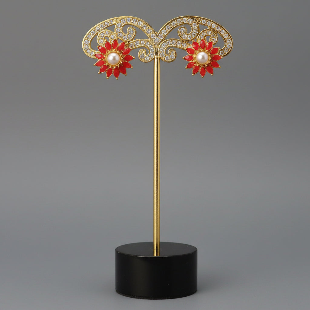 A pair of gold flower-shaped stud earrings with red enamel petals and white pearl centers, displayed on a wooden block stand.
