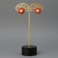 A pair of gold flower-shaped stud earrings with red enamel petals and white pearl centers, displayed on a wooden block stand.