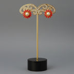 A pair of gold flower-shaped stud earrings with red enamel petals and white pearl centers, displayed on a wooden block stand.
