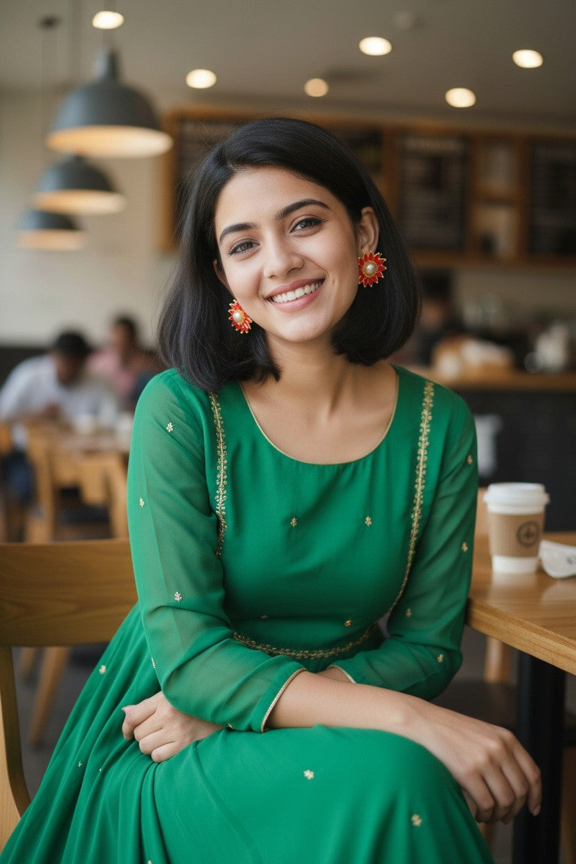 A woman with a short black bob haircut smiling while wearing a green long-sleeve dress with gold embroidery, seated in a cafe setting.