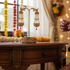 A close-up lifestyle shot of a person in a red and gold traditional Indian bridal outfit holding a gold pendant, with ornate gold earrings displayed on a stand on a wooden table in the background.