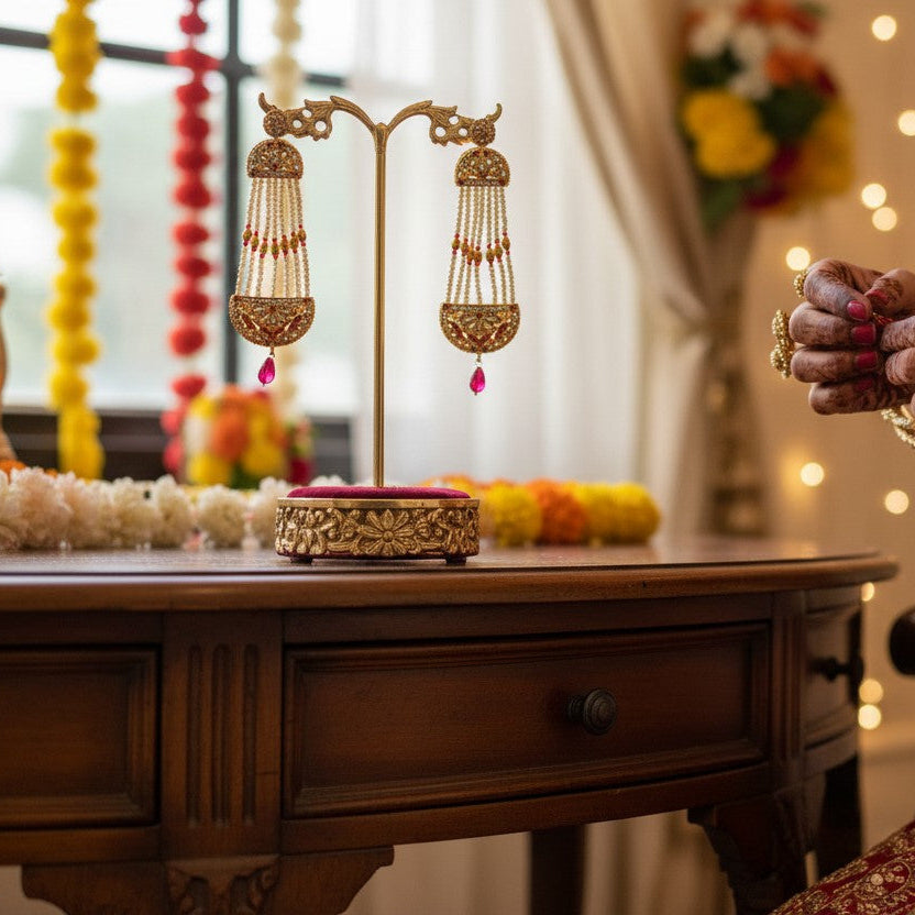 A close-up lifestyle shot of a person in a red and gold traditional Indian bridal outfit holding a gold pendant, with ornate gold earrings displayed on a stand on a wooden table in the background.