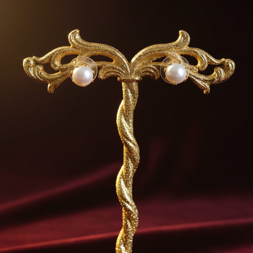 A pair of gold stud earrings with white pearl-like drops, displayed on a white wooden block stand against a red textured background.