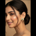 A close-up portrait of a woman wearing a square stud earring with a green center and a border of small clear stones, set against a warm beige background.