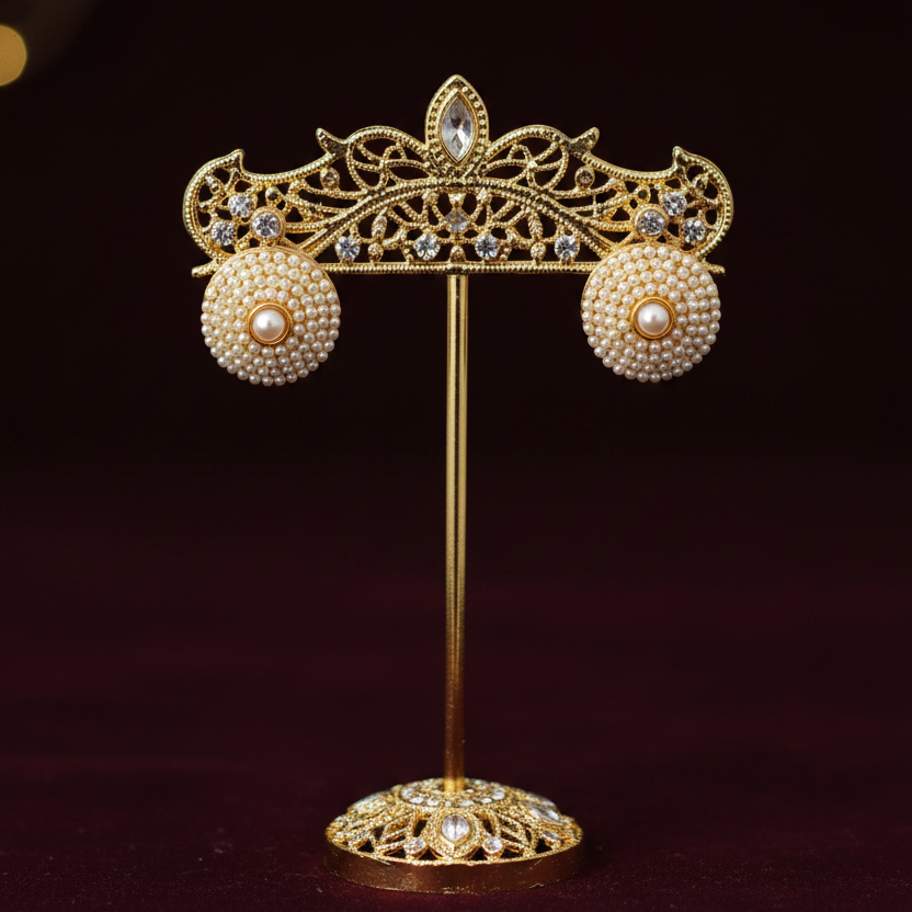 A pair of round gold-toned stud earrings decorated with small white faux pearls, displayed on a black stand atop a light wooden block with printed paper in the background.