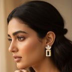 A close-up portrait of a woman wearing a dangling rectangular gold and white gemstone earring, with a matching ring on her finger, set against a soft beige background.
