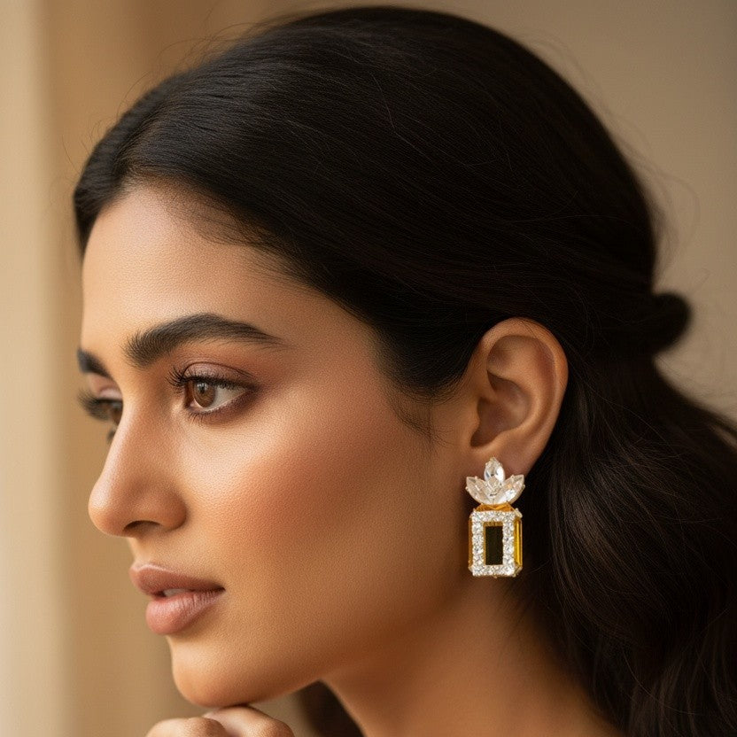 A close-up portrait of a woman wearing a dangling rectangular gold and white gemstone earring, with a matching ring on her finger, set against a soft beige background.