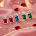 Four rectangular gemstone earrings in red, blue, and green, each surrounded by clear stones and topped with a leaf-like clear stone cluster, arranged on pink satin fabric with scattered rose petals and gold glitter.