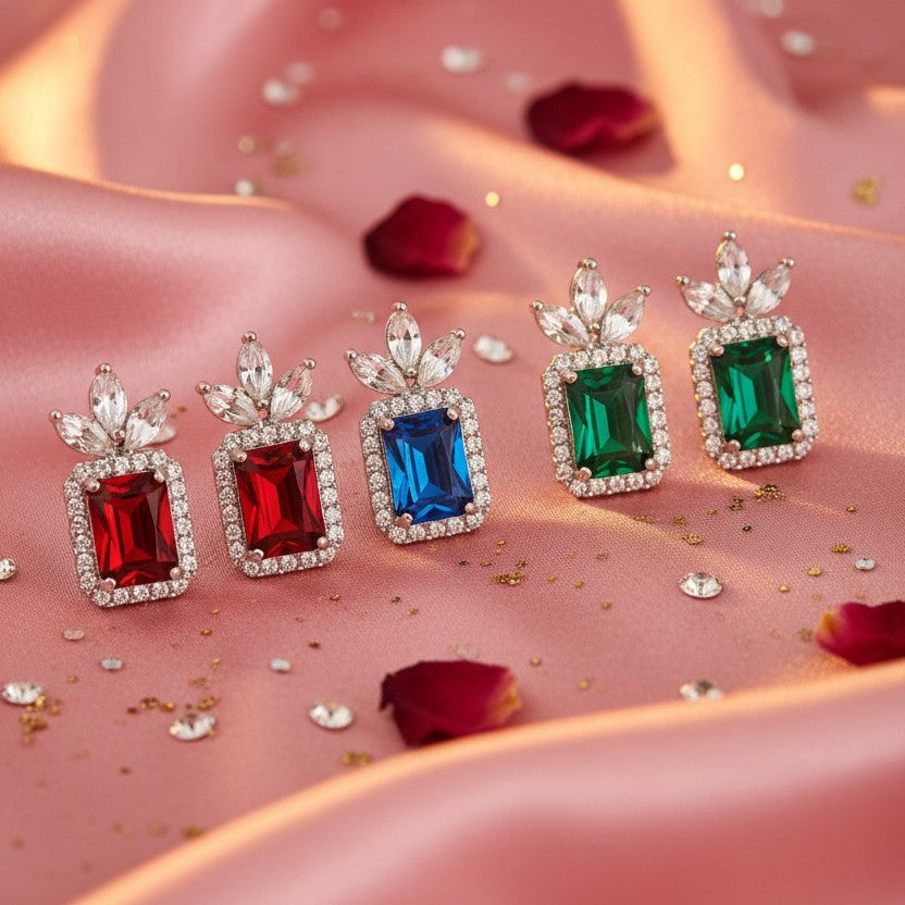 Four rectangular gemstone earrings in red, blue, and green, each surrounded by clear stones and topped with a leaf-like clear stone cluster, arranged on pink satin fabric with scattered rose petals and gold glitter.