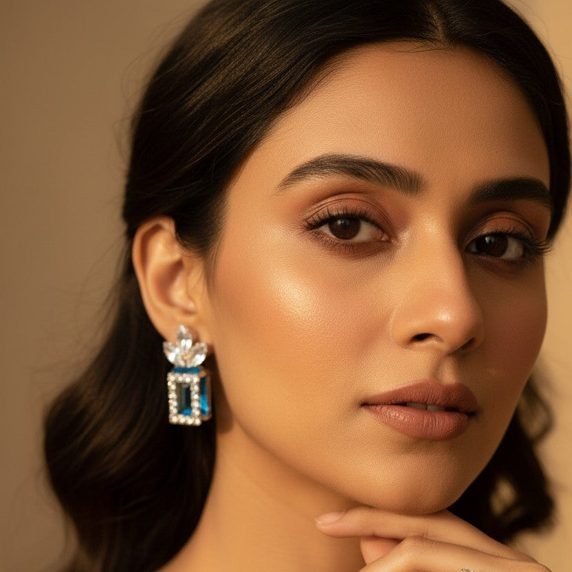 A close-up portrait of a woman wearing a dangling geometric earring with clear stones and blue accents, along with a simple ring on her finger, against a warm beige background.