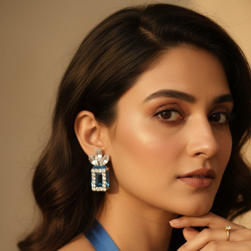 A close-up portrait of a woman wearing a dangling rectangular earring with clear stones and light blue accents, paired with a blue halter top against a warm beige background.