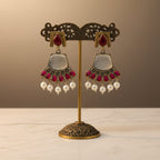 A pair of ornate gold and silver earrings with red stones and white pearl-like drops, held in a person's hand over a gray fabric surface.