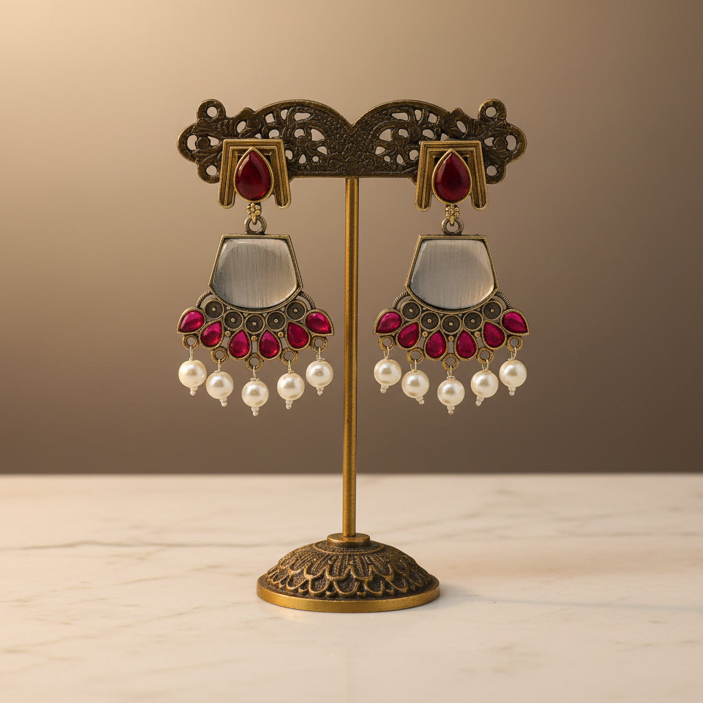 A pair of ornate gold and silver earrings with red stones and white pearl-like drops, held in a person's hand over a gray fabric surface.