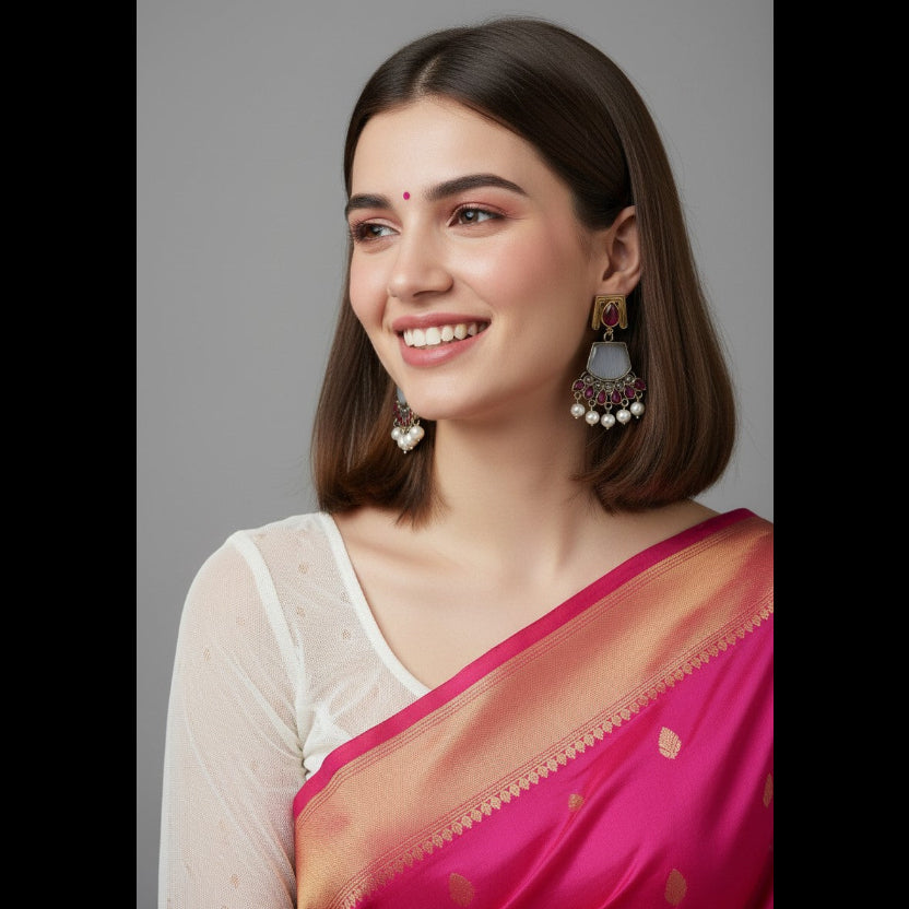 A woman with shoulder-length brown hair is shown in a close-up portrait, wearing a bright pink and peach-orange sari with a white blouse. She is wearing ornate dangling earrings and has a red bindi on her forehead, set against a solid gray studio background.
