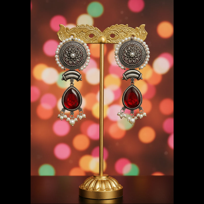 A pair of traditional Indian silver-toned dangle earrings with red teardrop stones and white pearl bead accents, displayed on a textured blue fabric surface.