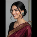A woman wearing a deep maroon silk saree with gold zari border detailing and a black blouse, accessorized with ornate silver earrings, shown in a close-up portrait.