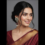 A woman with dark hair styled in an updo is shown in a close-up portrait, wearing a deep maroon sari with a gold border and matching maroon blouse. She is wearing ornate silver-toned earrings with red and white stones.