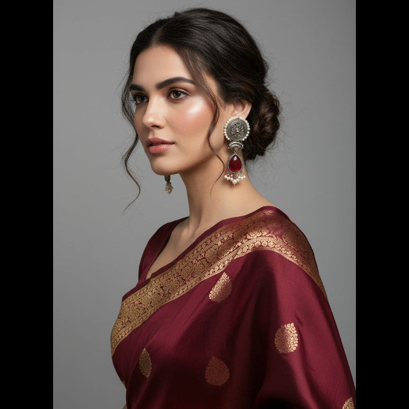 A woman wearing a deep maroon silk saree with gold zari embroidery and a wide gold border, paired with ornate silver and red earrings, photographed against a solid gray studio background.