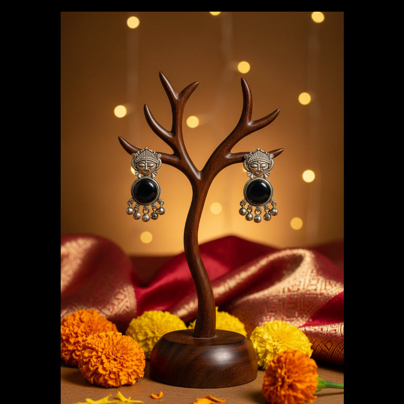 A pair of traditional silver-toned earrings with circular black stone centers and dangling silver bells, displayed on a textured gray fabric background.