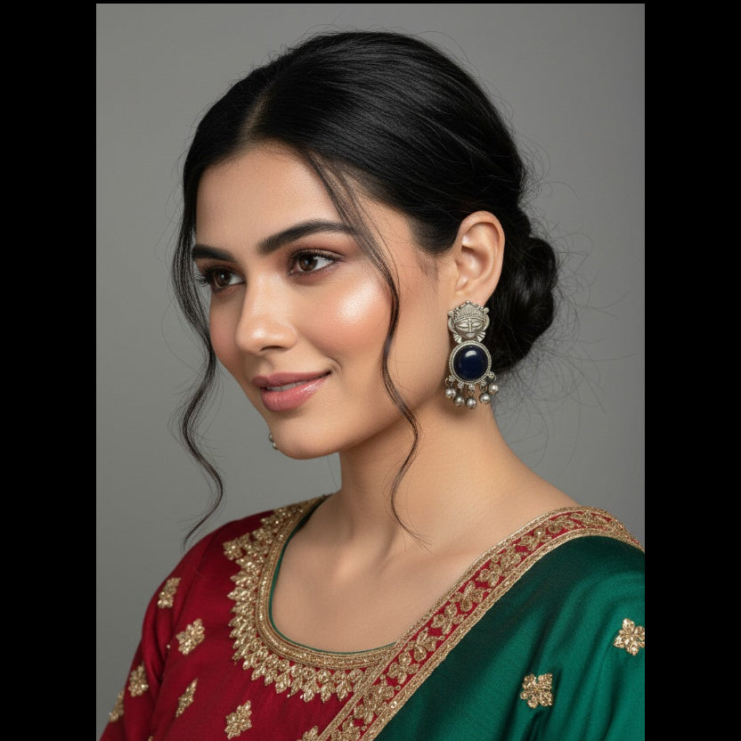 A woman with dark hair styled in a low bun is shown in a close-up portrait, wearing a deep green and maroon traditional outfit with gold embroidery and a matching earring with a dark blue stone.