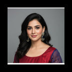 A woman with long, wavy black hair is shown from the chest up against a plain gray studio background. She is wearing a deep red top with sheer dark blue sleeves and large ornate silver-toned earrings.