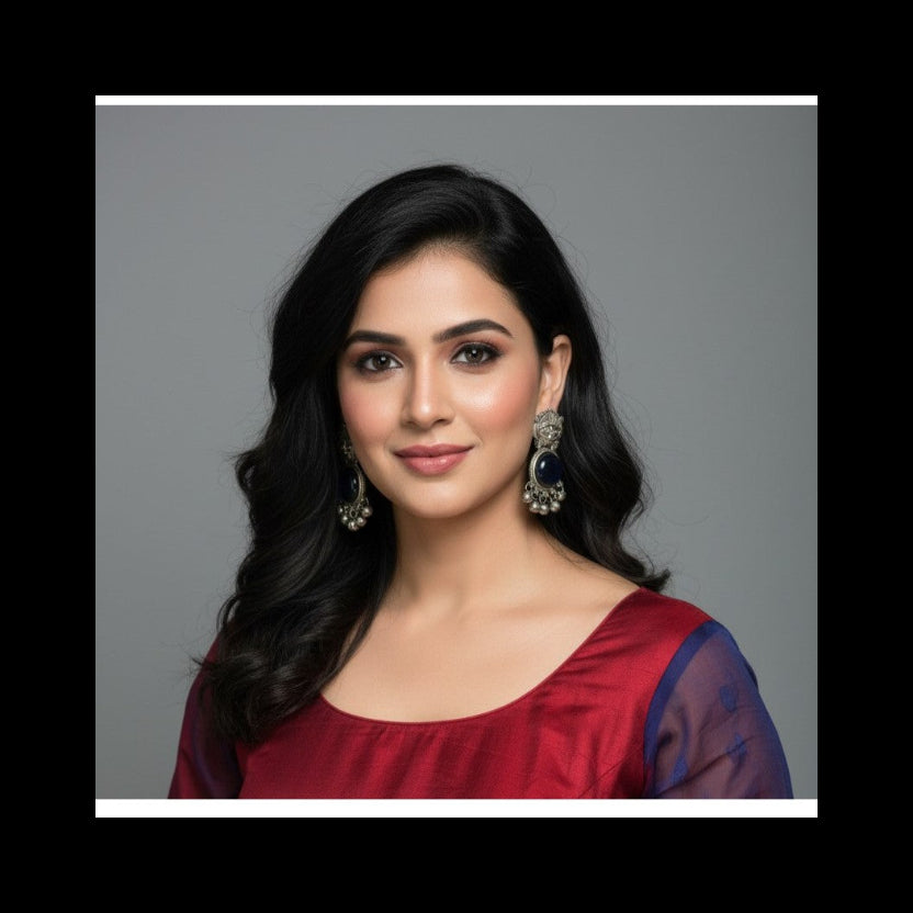 A woman with long, wavy black hair is shown from the chest up against a plain gray studio background. She is wearing a deep red top with sheer dark blue sleeves and large ornate silver-toned earrings.