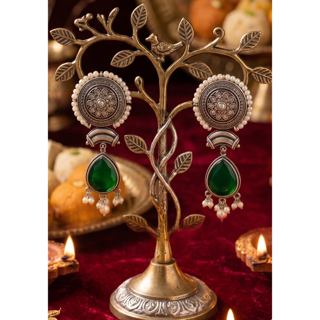 A pair of ornate silver-toned earrings with circular floral motifs, white pearl-like beads, and green teardrop drops, displayed on a decorative tree-shaped jewelry stand over a red fabric surface with warm lights in the background.