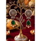 A pair of ornate silver-toned earrings with circular floral motifs, white pearl-like beads, and green teardrop drops, displayed on a decorative tree-shaped jewelry stand over a red fabric surface with warm lights in the background.