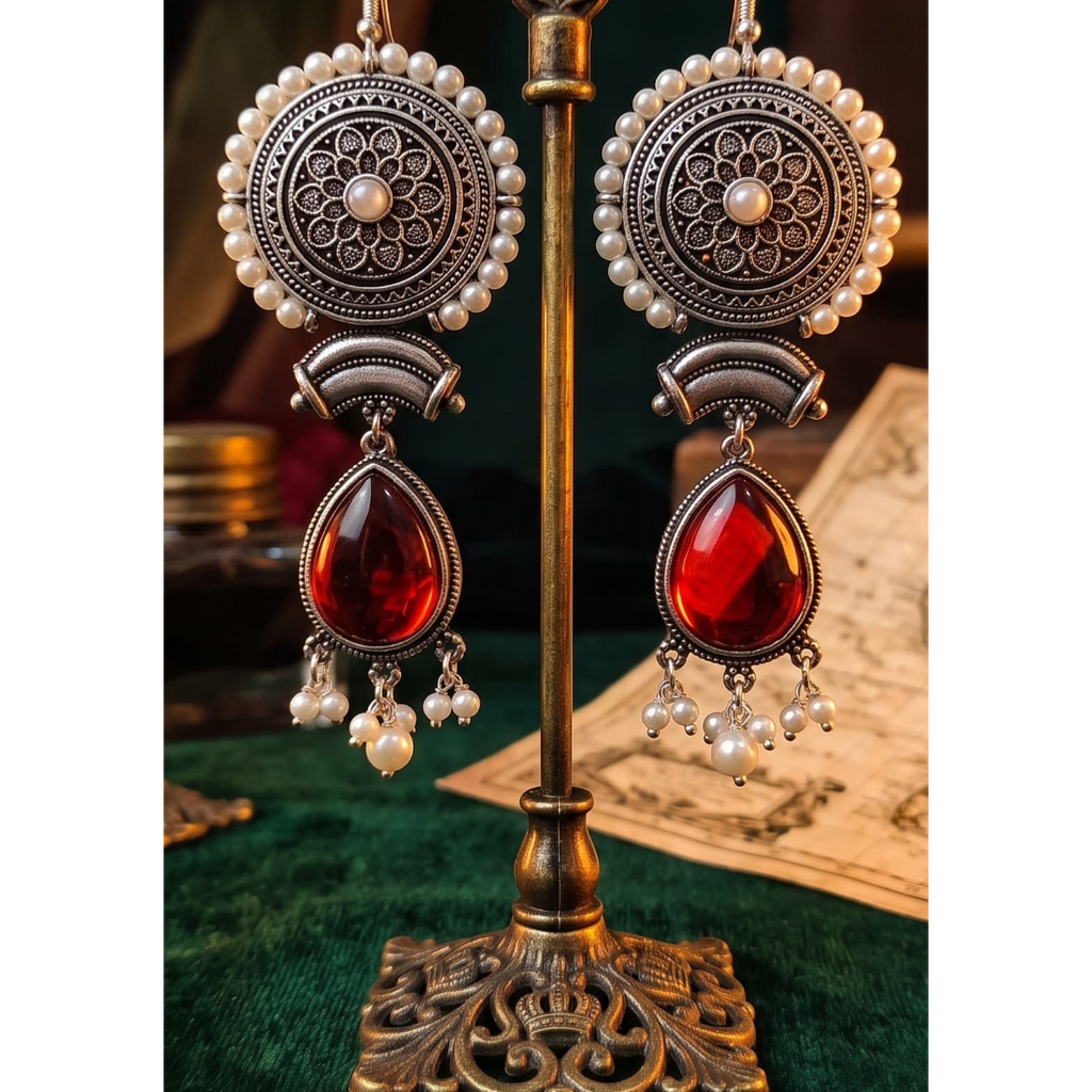 A pair of ornate dangling earrings displayed on a decorative metal stand. The earrings feature circular silver-toned filigree motifs with white pearl-like beads and large teardrop-shaped deep red gemstones hanging below.