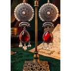 A pair of ornate dangling earrings displayed on a decorative metal stand. The earrings feature circular silver-toned filigree motifs with white pearl-like beads and large teardrop-shaped deep red gemstones hanging below.