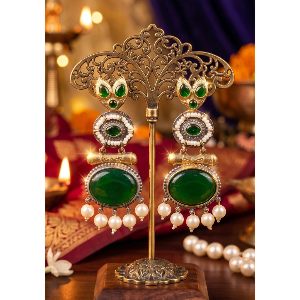 A pair of ornate gold-toned dangle earrings with large green stone centers, smaller green and white stones, and dangling pearl-like beads, displayed on a decorative stand in a warm, richly lit setting.
