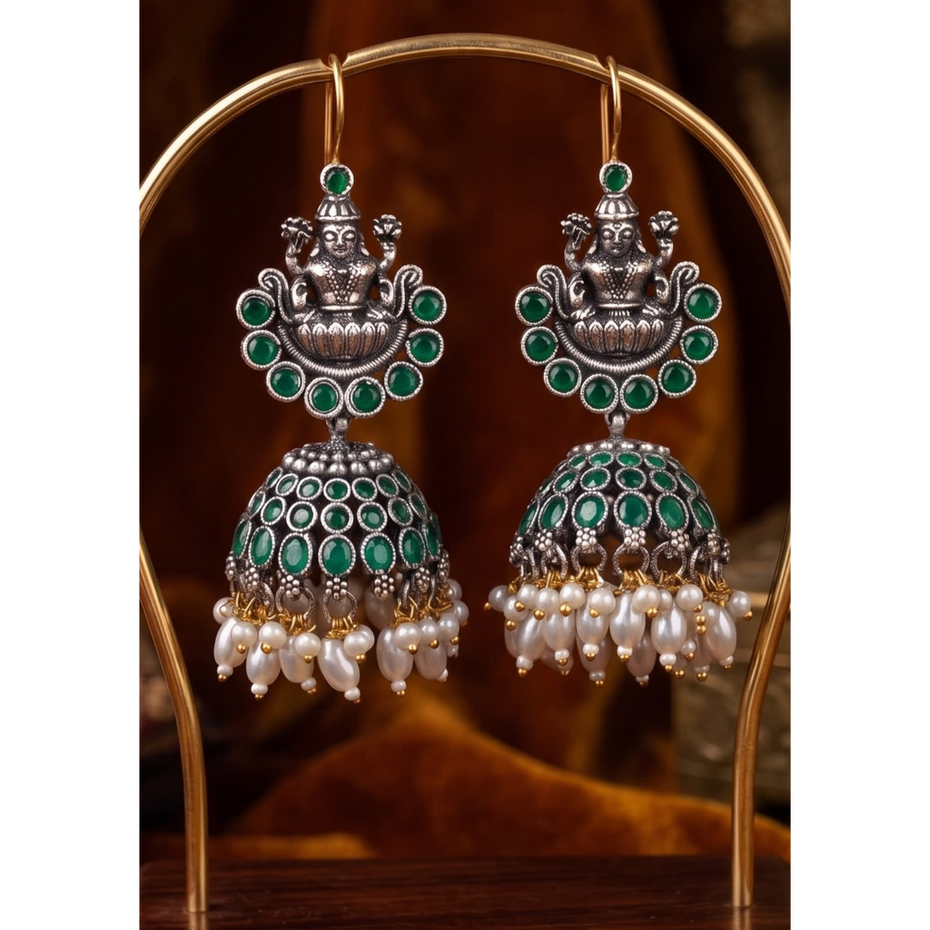 A pair of traditional Indian silver-toned jhumka earrings with green stone accents and white pearl bead drops, displayed on a gold-colored stand against a warm brown background.