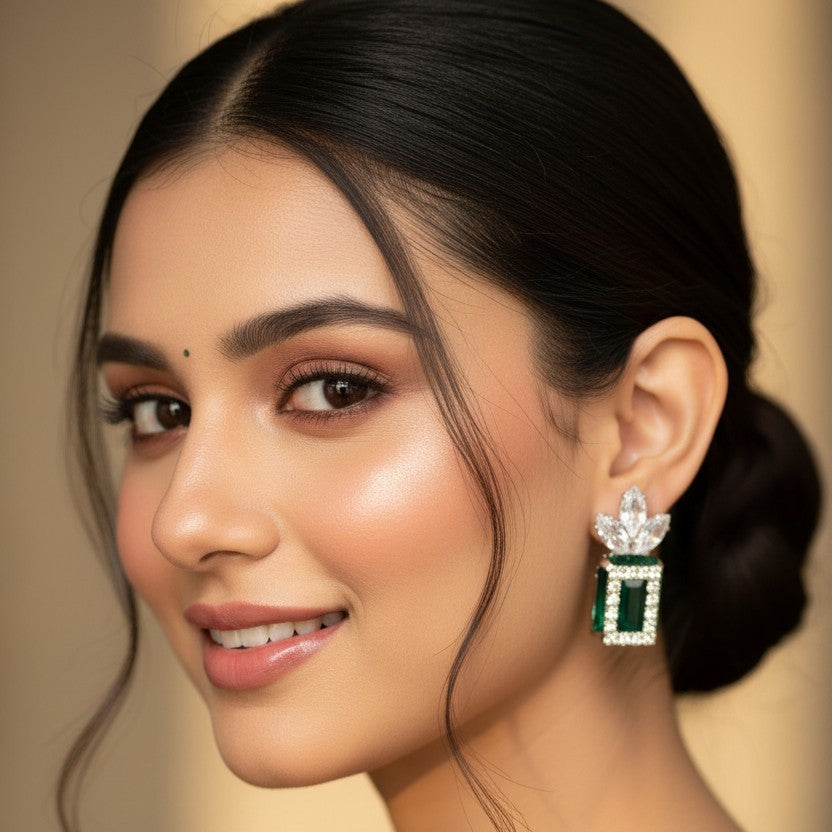 A close-up portrait of a woman wearing a dangling earring with a silver-toned floral top and a rectangular green stone setting. The background is a soft, warm beige studio backdrop.