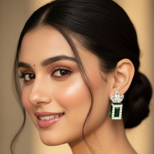 A close-up portrait of a woman wearing a dangling earring with a silver-toned floral top and a rectangular green stone setting. The background is a soft, warm beige studio backdrop.