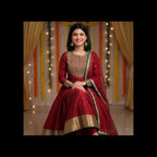 A woman wearing a traditional red Indian salwar kameez outfit with gold embroidery and a green-trimmed dupatta, posed against a decorated backdrop with gold curtains and string lights.