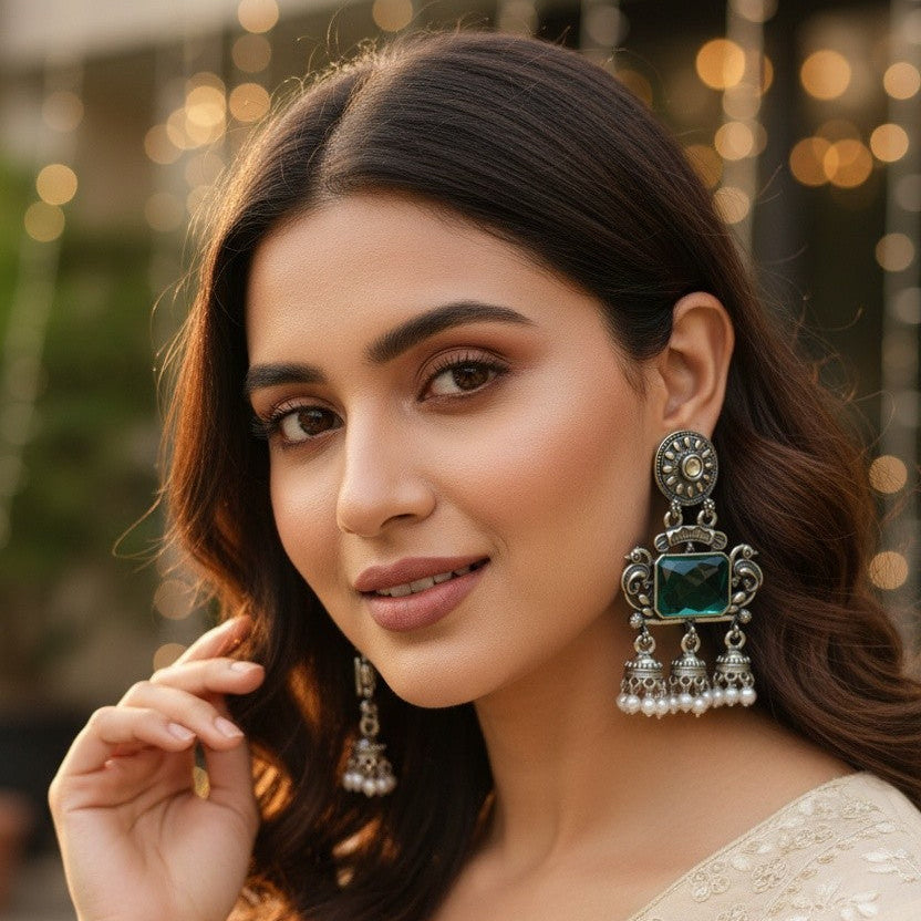 A woman wearing ornate silver-toned traditional earrings with green stones and dangling pearl-like accents, paired with a beige embroidered top. The background is softly blurred with warm bokeh lights.