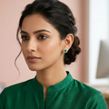 A woman with dark hair styled in a low bun wears a vibrant green high-neck top and a matching green drop earring, set against a soft-focus office background with pink walls.