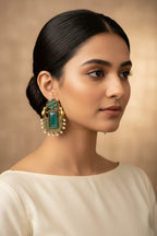 A pair of ornate gold-toned earrings with rectangular green turquoise-like stones and small bead details, displayed on a textured gray fabric background.